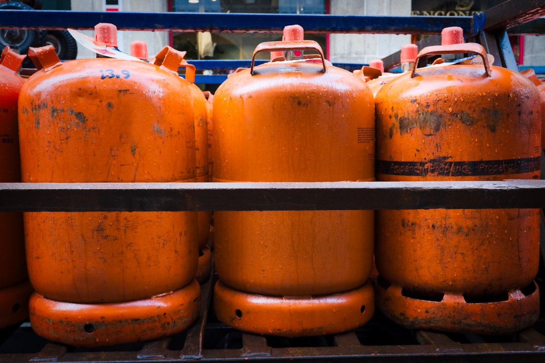 Orange Propane Tanks Stacked on Metal Rack in City Setting - A close-up shot of three large orange propane tanks stacked on a metal rack in an urban setting. The tanks are in a row, and the rack appears to be part of a truck or loading dock. The tanks are worn and faded with evidence of rust and water droplets.