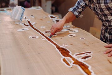 Carpenter cleaning the wooden table from epoxy resin drops in a workshop. Process of making a craft resin and wood table