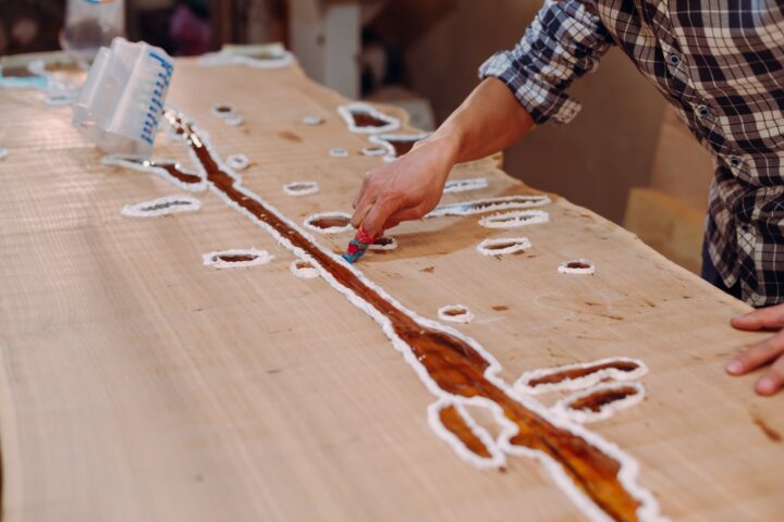 Carpenter cleaning the wooden table from epoxy resin drops in a workshop. Process of making a craft resin and wood table